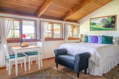 Cozy bedroom with a bed, dining table, and a black chair, featuring wooden beams and large windows.