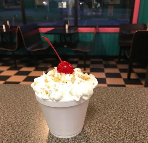A cup of ice cream topped with whipped cream, crushed nuts, and a cherry, set on a checkered table in a diner.