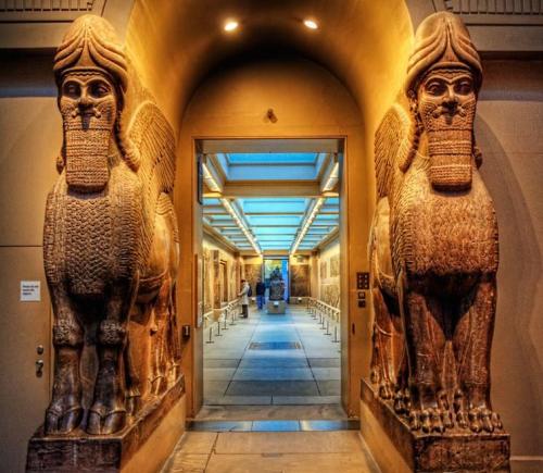 Two large stone winged bulls stand at the entrance of a museum hallway, leading to an illuminated exhibit.