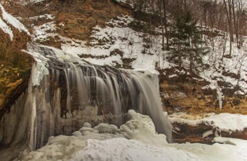 A frozen waterfall cascading over rocky cliffs, surrounded by snow-covered trees and icy terrain.