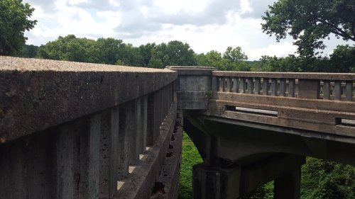 A close-up view of a concrete bridge railing with greenery and trees in the background under a cloudy sky.