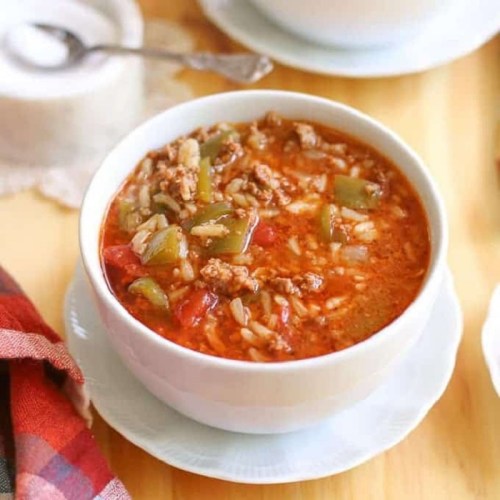 A bowl of hearty soup with ground meat, rice, green peppers, and tomatoes, served on a wooden table.