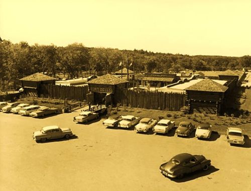 Historic fort with wooden structures and vintage cars parked in front, surrounded by trees and open land.