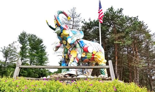Colorful elephant sculpture made of plates, surrounded by flowers and trees, with an American flag in the background.