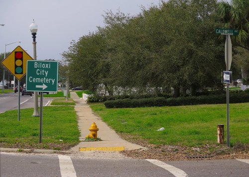 Signpost directing to Biloxi City Cemetery, with a traffic light and a fire hydrant nearby, on a grassy corner.