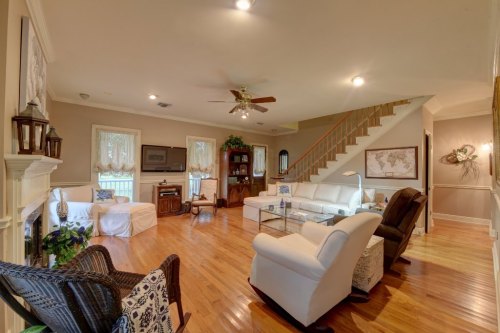 Spacious living room with hardwood floors, white furniture, a ceiling fan, and a staircase in the background.
