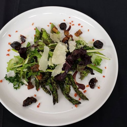 A vibrant salad with asparagus, mixed greens, shaved cheese, and dark berries, garnished with red pepper flakes.