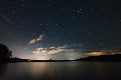 A serene night sky filled with stars over a calm lake, with a few clouds and shooting stars visible.