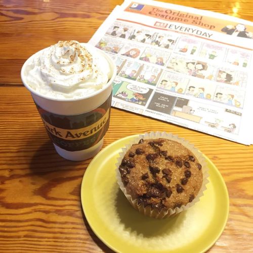 A cup of whipped cream-topped coffee next to a chocolate chip muffin on a yellow plate, with a newspaper in the background.