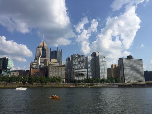 A view of a city skyline with tall buildings and a river, featuring a kayak on the water under a partly cloudy sky.