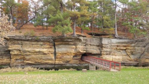 A wooden bridge leads to a rock formation with a cave, surrounded by trees in autumn colors.