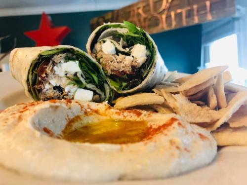 A plate featuring two wraps filled with greens and chicken, served with hummus drizzled with olive oil and pita chips.