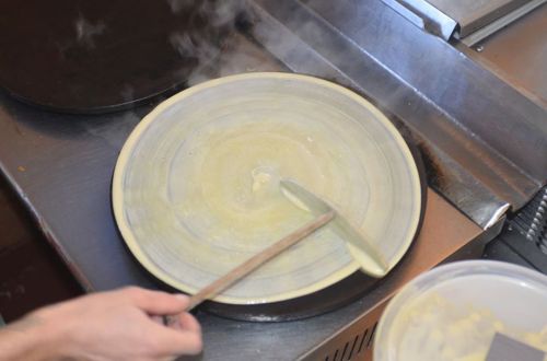 A hand spreads batter on a hot griddle, creating a thin layer for a crepe or pancake. Steam rises from the surface.