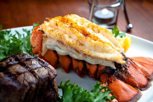 A gourmet plate featuring grilled lobster tail and a steak, garnished with lettuce and a lemon wedge.