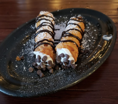 Two cannoli filled with cream, topped with chocolate drizzle and dusted with powdered sugar, served on a black plate.