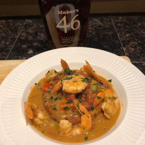 A plate of shrimp and sauce served over a piece of meat, with a bottle of Maker's 46 bourbon in the background.
