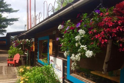 A rustic building with colorful flower baskets and a sign reading "The Deck," surrounded by greenery and wooden decor.