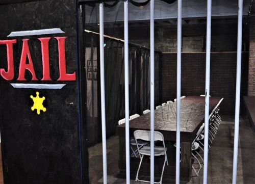 A dimly lit room with a long table and chairs, behind jail bars, featuring a sign that reads "JAIL."