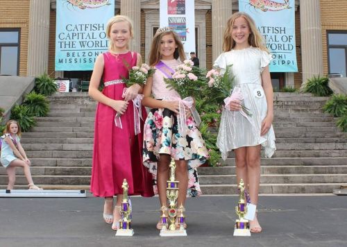 Three girls in formal dresses hold flowers and trophies, smiling on stage at a pageant in Belzoni, Mississippi.