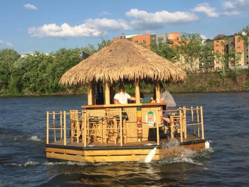 A floating tiki bar with a thatched roof on a river, surrounded by greenery and buildings in the background.