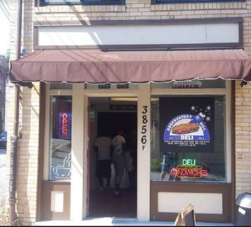 A small deli with a brown awning, neon "OPEN" sign, and a menu displayed in the window. People are entering.
