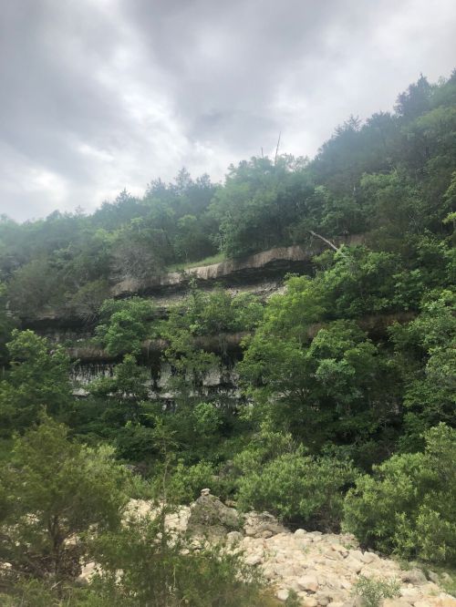 Lush green hillside with rocky layers under a cloudy sky, featuring trees and shrubs along a riverbed.