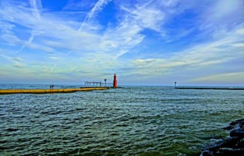 A serene seascape featuring a red lighthouse, a pier, and a calm blue sky with wispy clouds over the water.