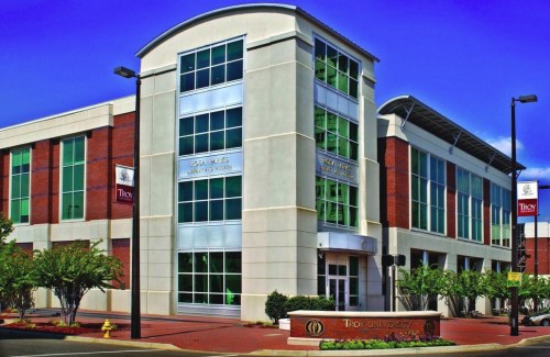 Modern building of Troy University with large glass windows and a welcoming entrance, surrounded by greenery.