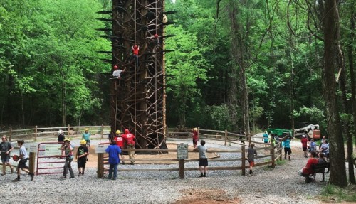 A climbing tower surrounded by trees, with people climbing and others watching or resting nearby.