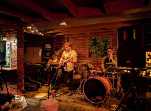 A band performs in a cozy venue with brick walls, featuring a guitarist and a drummer surrounded by warm lighting.