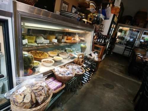 A cozy shop interior featuring a display case with various foods, baskets of baked goods, and shelves of wine.