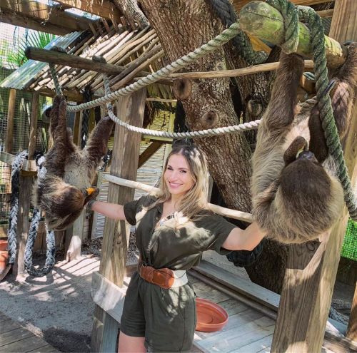 A woman in a green outfit smiles while holding onto ropes with two sloths hanging nearby in a natural setting.