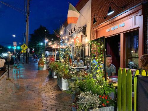 A lively street scene at night featuring an Irish pub with outdoor seating, string lights, and colorful plants.