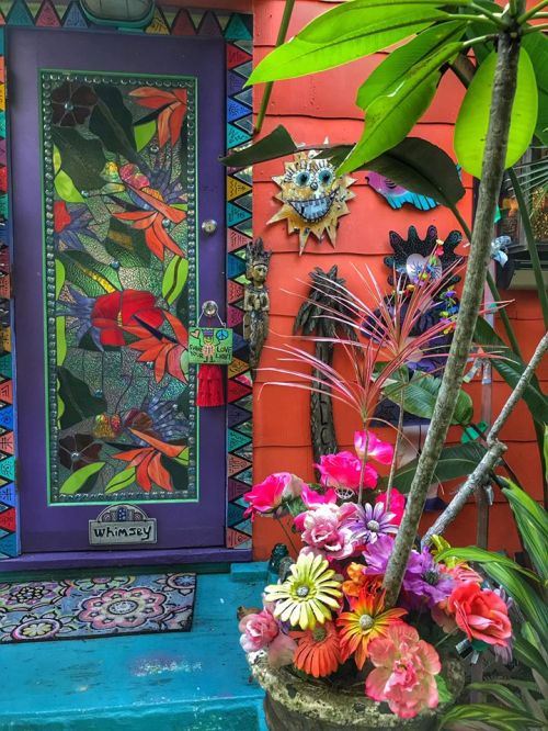 Colorful door adorned with vibrant flowers and artwork, surrounded by lush greenery and a decorative planter.