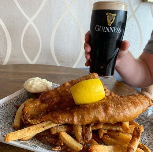 A plate of fish and chips with a lemon slice, accompanied by a pint of Guinness and a small bowl of sauce.