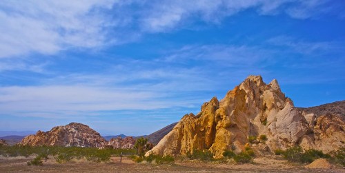 A scenic view of rugged rock formations under a blue sky with wispy clouds, surrounded by desert vegetation.