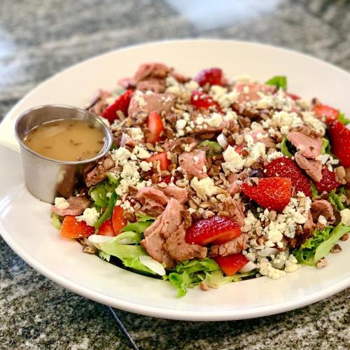 A colorful salad with greens, strawberries, pecans, and feta cheese, served with a small cup of dressing on the side.
