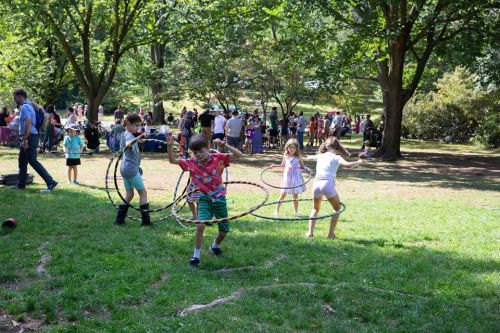 Children playing with hula hoops in a park, surrounded by trees and a crowd of people enjoying the day.
