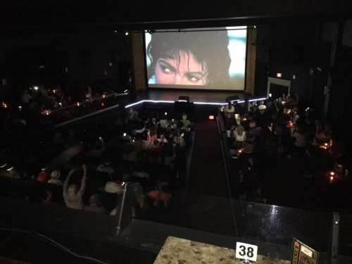 A dimly lit theater filled with an audience watching a film on a large screen, with candles on tables.