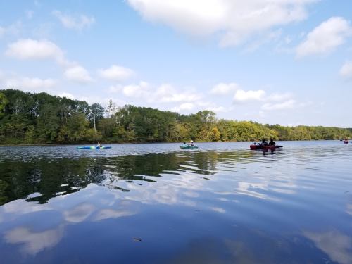 A serene lake scene with kayakers paddling on calm waters, surrounded by lush green trees and a blue sky.