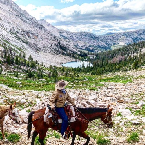 A person on horseback in a mountainous landscape, with a lake and lush greenery in the background.