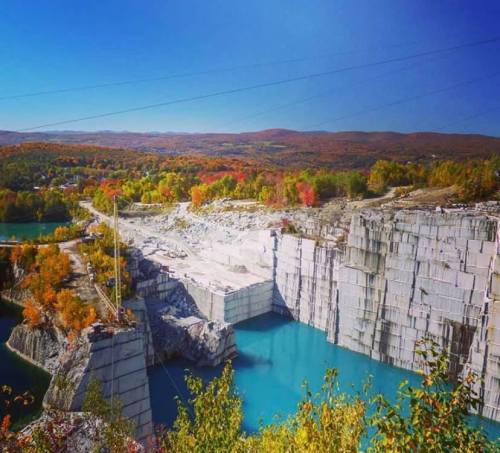 A scenic view of a quarry with blue water, surrounded by colorful autumn foliage and clear blue skies.