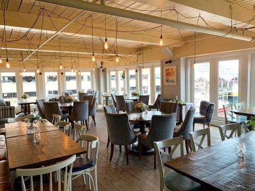 Brightly lit restaurant interior with wooden tables, chairs, and large windows overlooking a waterfront view.