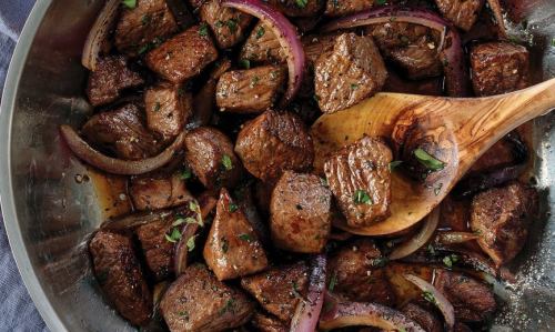 A close-up of sautéed beef cubes with red onions and herbs in a silver pan, with a wooden spoon for serving.