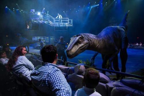 A lifelike dinosaur interacts with an audience at a live show, with performers in the background and dramatic lighting.