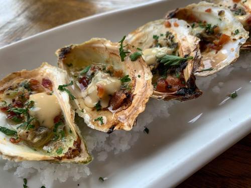 Grilled oysters topped with herbs and sauce, served on a bed of salt on a white plate.