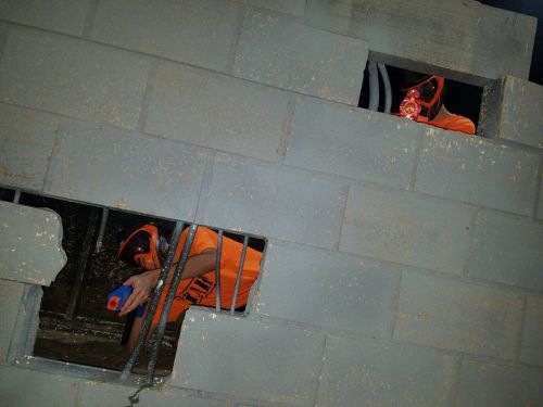 Two individuals in orange shirts and masks are peering through openings in a gray concrete wall.
