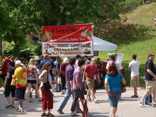 A busy outdoor festival scene with people walking past a colorful food stall offering Mexican cuisine and drinks.