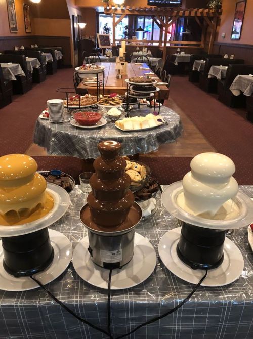 A buffet setup featuring three chocolate fountains (yellow, brown, and white) surrounded by various desserts and snacks.