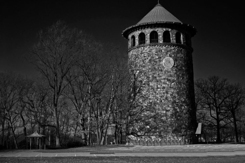 A tall, stone tower with a clock, surrounded by bare trees, set against a dark sky in black and white.
